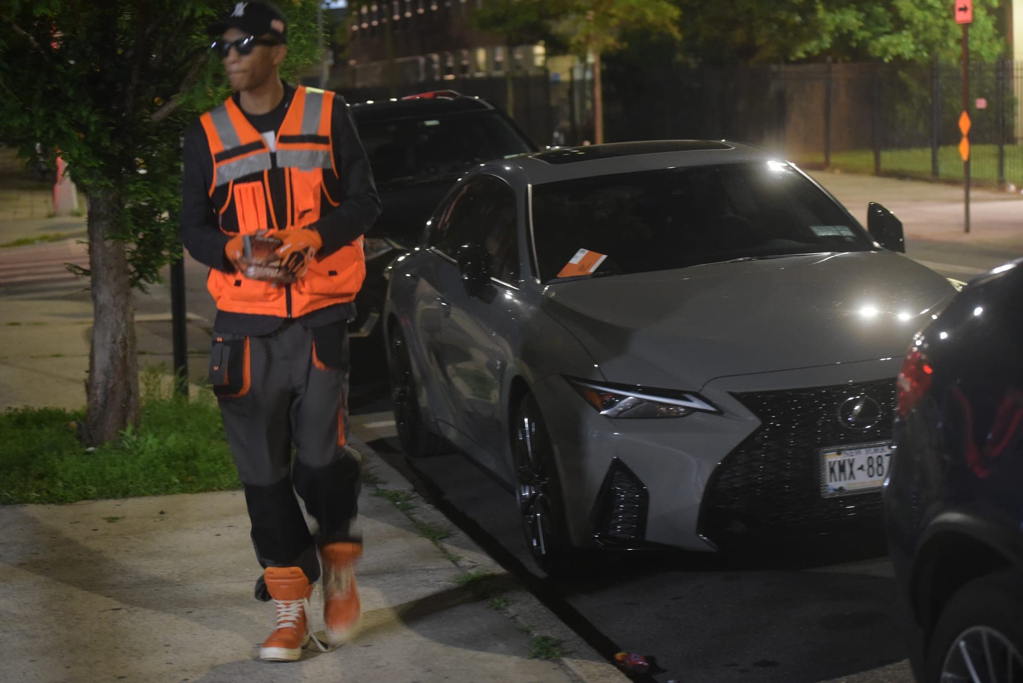 SNVRK in hi-vis workwear on a sidewalk beside parked cars at night
